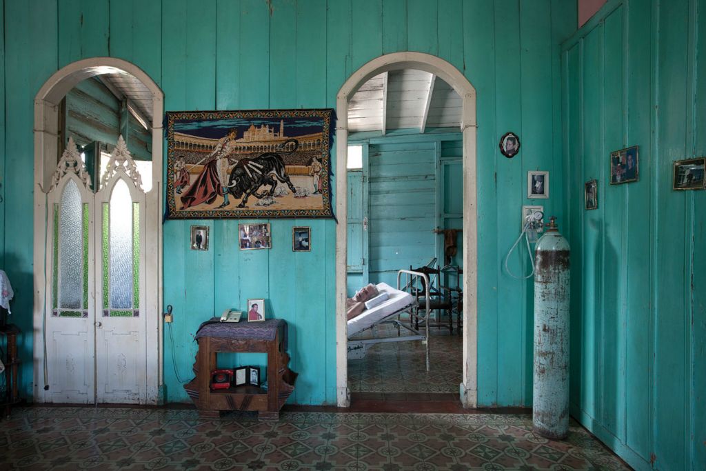 Vincente suffers from dementia and is cared for by his daugher who is also caring for her bedridden mother. She has placed Vicente in the doorway so he can still be a part of famly life.